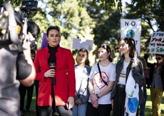 A television journalist reports live from a school climate strike in Melbourne as a camera operator films